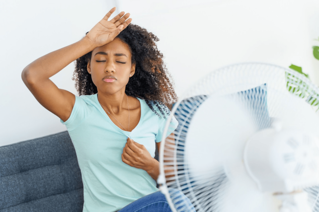 A young woman is sitting in front of a fan. Her hand is against her forehead, suggesting she is hot.